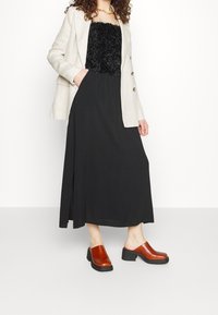 Zwarte maxi-rok gecombineerd met een zwart topje met structuur en een licht crèmekleurig blazer, aangevuld met bruine klompen met hak.