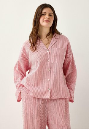 Ung kvinde iført løs rød- og hvidstribet skjorte med knapper og matchende bukser, stående med hænderne i lommerne mod en ensfarvet baggrund.