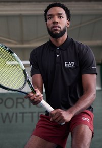 Schwarzes Polo-Shirt mit EA7-Logo, rote Shorts und ein Tennisschläger mit grünem und schwarzem Design. Indoor-Sportumgebung mit grünem Hintergrund.