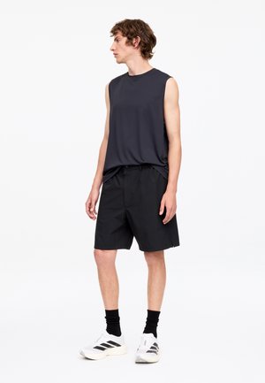 Giovane uomo rivolto verso sinistra che indossa una canotta nera senza maniche, pantaloncini neri, calzini neri e scarpe da ginnastica bianche con strisce nere.