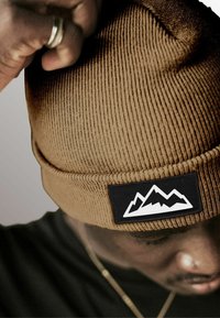 Brown ribbed beanie with a black rectangular patch featuring a white mountain graphic. Soft textile with a snug fit.