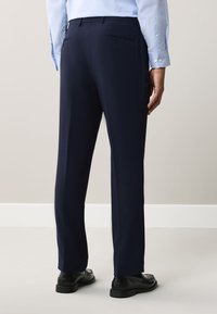Uomo che indossa pantaloni formali blu scuro e scarpe eleganti nere, mostrato di spalle su uno sfondo beige uniforme.
