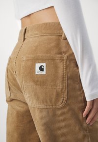 Salopette en velours côtelé marron avec une finition texturée et un patch logo Carhartt sur la poche arrière, associée à un t-shirt ajusté à manches longues blanc.