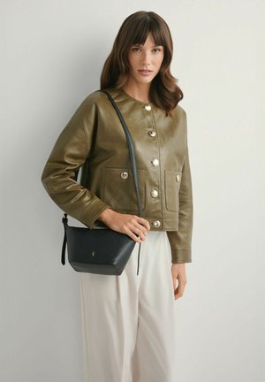 Kobieta ubrana w oliwkowo-zieloną skórzaną kurtkę z guzikami i białe spodnie, trzymająca czarną torebkę na ramię ze złotym logo na tle szarej ściany.