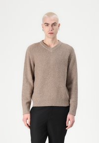 Beige knitted sweater with a V-neck, textured pattern, and ribbed hems, paired with black trousers. No other details present.