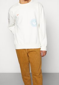 Felpa in cotone bianco sporco con scollo rotondo, caratterizzata da una grafica di sole blu sul davanti e un logo arancione sul petto.