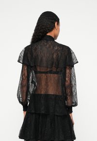 Zwarte kanten blouse met een doorzichtig rugpand, gelaagd ontwerp, volumineuze mouwen en een hoge kraag. Gelaagd kant met bloemenpatronen.