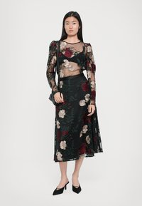 Zwarte jurk met bloemenborduursel in rood en beige, doorzichtige lange mouwen, een aansluitend lijfje en een vloeiende rok, gecombineerd met zwarte hakken.