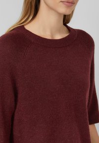 Pull en tricot bordeaux avec un col rond côtelé et des manches courtes, présentant une texture douce et une coupe légèrement ample.