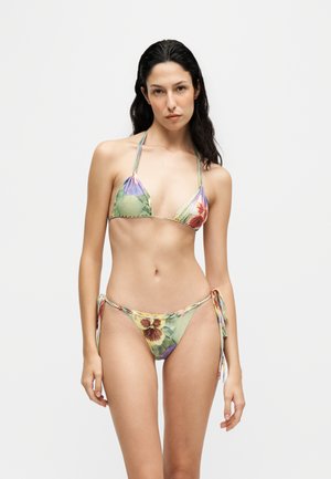 Kvinde med langt mørkt hår iført en grøn bikini med blomsterprint og bindebånd, stående mod en ensfarvet lys baggrund.