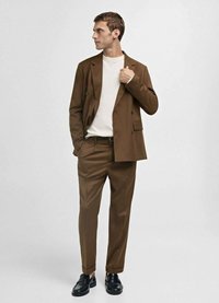Bruine op maat gemaakte colbert over een wit overhemd, gecombineerd met bijpassende bruine broek en zwarte instappers. Glad materiaal, moderne snit.