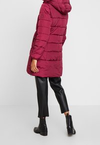 Burgunderfarbene Daunenjacke mit Kapuze, mit einem gesteppten Design, Fronttaschen und einem Saum, der über das Knie reicht.