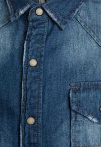 Camisa de mezclilla con un diseño clásico, color azul, costuras contrastantes, botones a presión y un bolsillo en el pecho con bordes deshilachados.
