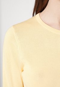 Lys gul strikket sweater med rund halsudskæring, ribbede manchetter og glat tekstur. Nærbillede fokuserer på stofdetaljer og farve.