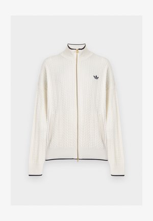 Mann trägt einen weißen, strukturierten Zip-up-Pullover mit schwarzem Logo auf der linken Brust, kombiniert mit schwarzen Hosen, steht vor einem einfarbigen Hintergrund.