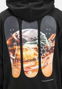Čierna mikina s kapucňou, ktorá na zadnej strane obsahuje veľkú grafiku zobrazenú skateboardistu. Grafika obsahuje živé farby a dynamické prvky pohybu.