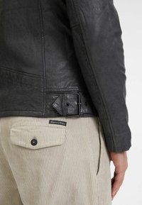 Giubbotto di pelle nera con dettaglio di fibbia laterale, indossato sopra pantaloni di corduroy beige chiaro con una tasca con bottone e etichetta del marchio.