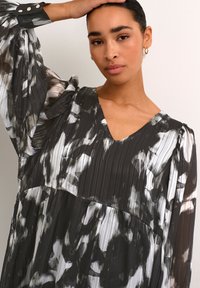 Robe plissée à motifs noir et blanc avec un décolleté en V, des manches longues bouffantes et des accents de boutons sur les poignets. Tissu lisse et fluide.
