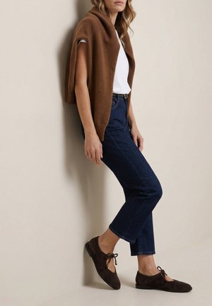 Vrouw draagt donkerblauwe spijkerbroek, wit t-shirt, bruine trui over haar schouders gedrapeerd en donkerbruine suède loafers met strikdetails.