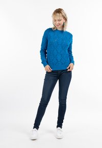 Kék kötött pulóver, gyémántmintával és texturált részletekkel, sötétkék skinny farmerral és fehér sportcipővel párosítva.