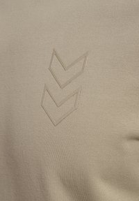 Beige sweatshirt tyg med en texturerad yta, med en upphöjd, ljusbeige chevronslogotyp på vänster sida.