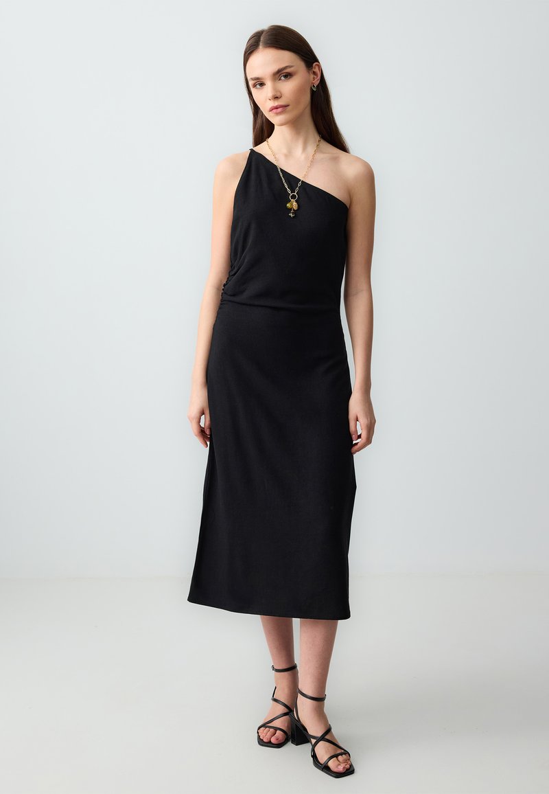 Zwarte, mouwloze jurk met een asymmetrische halslijn en geplooide details bij de taille. Gecombineerd met zwarte sandaaltjes met banden.
