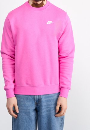 Person wearing a bright pink Nike sweatshirt and blue jeans, standing with hands relaxed at sides against a plain background.