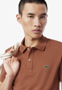 Brązowa koszulka polo z teksturowanej dzianiny, z dwoma guzikami przy plastrze i haftowanym logo krokodyla na lewej piersi.