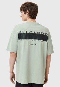 Camiseta de gran tamaño verde claro con un panel horizontal negro y texto en la parte trasera. Material de algodón suave, corte relajado.