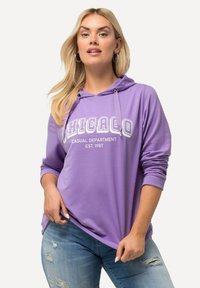 Felpa viola con il testo "CHICAGO", design casual, maniche lunghe e cappuccio con cordino. Indossata sopra jeans blu strappati.