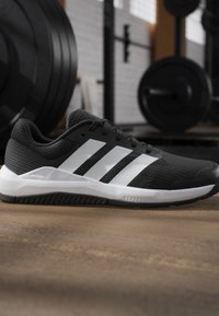 Chaussure de sport noire et blanche avec trois bandes blanches sur le côté, posée sur le sol d’une salle de sport avec du matériel de musculation en arrière-plan.