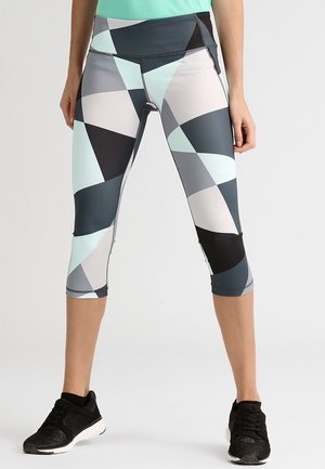 Nogi w wielokolorowych leginsach typu capri z geometrycznym wzorem i czarnych butach sportowych na jednolitym jasnym tle.