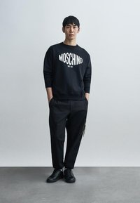 Μαύρο φούτερ Moschino με λευκό λογότυπο, συνδυασμένο με άνετο μαύρο παντελόνι με τσέπη φερμουάρ και μαύρα αθλητικά παπούτσια, με απλό φόντο.