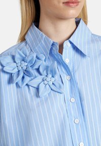 Camisa de rayas azul claro con dos flores de tela en el lado izquierdo, con pétalos plisados y detalles de botones en la parte delantera.
