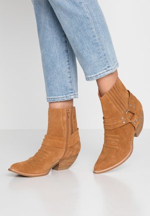 Jasnoniebieskie skrócone dżinsy w połączeniu z beżowymi zamszowymi botkami za kostkę, wyposażonymi w boczne zamki, ozdobne paski oraz niskie, ułożone na stosy obcasy.