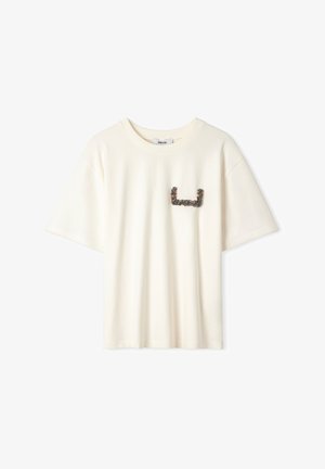 Kremowa bawełniana koszulka z krótkim rękawem, z teksturowaną grafiką w ciemnym odcieniu po lewej stronie klatki piersiowej. Miękki, luźny krój.