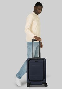 Giovane uomo con camicia beige a maniche lunghe e jeans che trascina una valigia rigida blu scuro con manico esteso e quattro ruote su sfondo grigio.