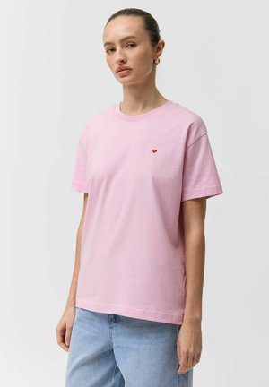 Kobieta o jasnej karnacji i związanych do tyłu włosach ubrana jest w różowy T-shirt z małym czerwonym sercem na piersi, w połączeniu z jasnoniebieskimi dżinsami.