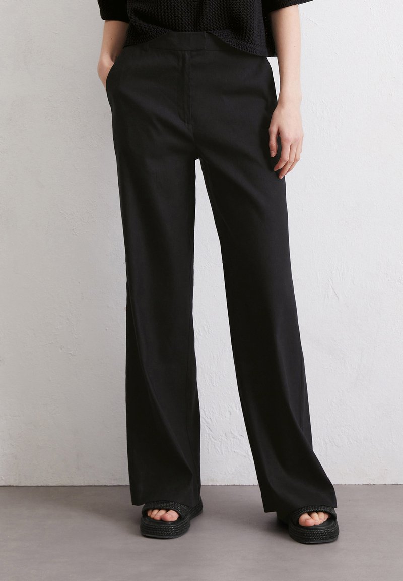 Zwarte wijde broek van een soepele stof met een hoge taille. Heeft een gulp aan de voorkant en zijzakken, gedragen met stevige sandalen.