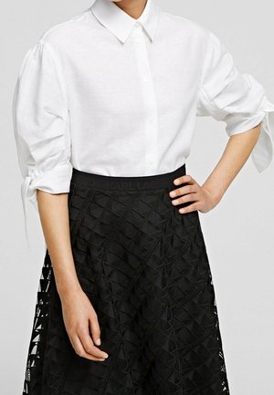 Femme portant une chemise blanche boutonnée avec des manches bouffantes attachées aux coudes et une jupe noire taille haute avec des motifs géométriques.