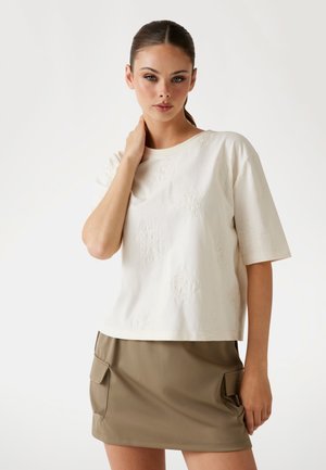 Cream cotton T-shirt with embossed patterns, round neckline, loose fit; paired with a khaki mini skirt featuring side pockets and smooth texture.