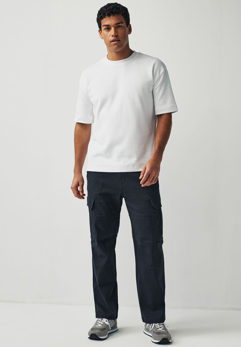 Weißes Baumwoll-T-Shirt mit kurzen Ärmeln, kombiniert mit dunklen Cargo-Hosen mit Seitentaschen und grauen Sportschuhen. Einfaches, lässiges Outfit.