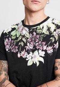 Person som bär en svart t-shirt med ett färgglatt blommönster som har rosa och vita blommor samt gröna blad över bröstet och axlarna.