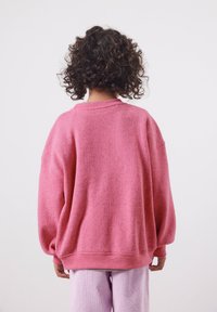Personne aux cheveux bouclés et foncés, portant un pull rose ample et un pantalon violet clair, debout face à un fond uni clair.