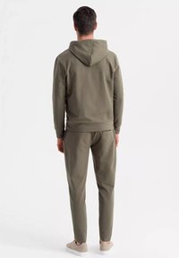 Conjunto de sudadera verde oliva y pantalones a juego, hecho de tela suave, con capucha ajustable y puños acanalados. Modelo visto desde la espalda.