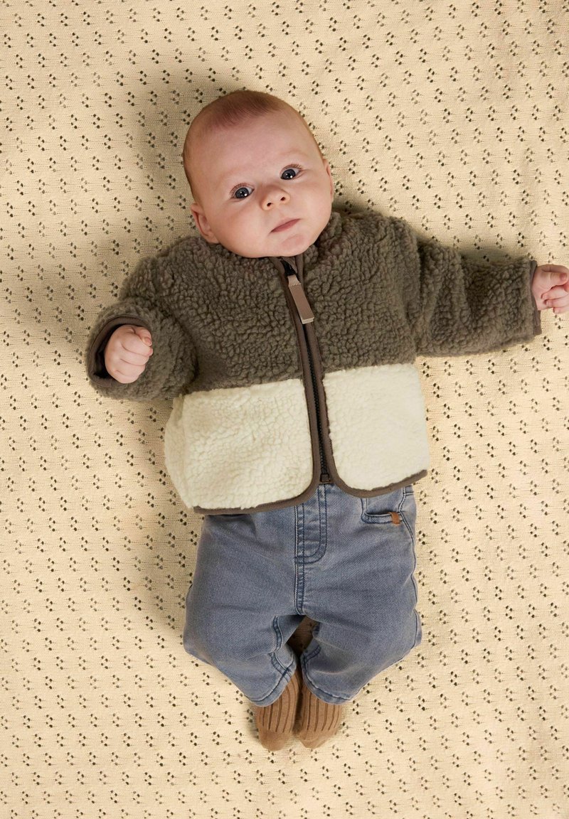 Bambino con indosso una giacca in pile marrone e crema, pantaloni blu e calzini marroni, sdraiato su una coperta beige con motivo, che guarda verso l'alto.