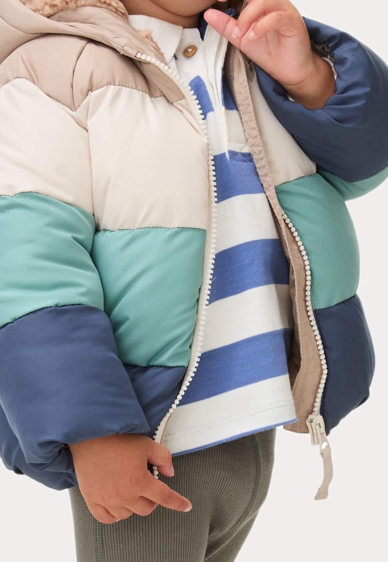 Enfant portant une doudoune color block beige, sarcelle, marine et crème sur une chemise rayée bleu et blanc.