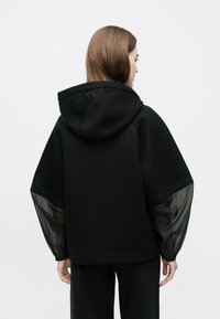 Črna jopa s teksturirano tkanino, kontrastnimi prosojnimi rokavi in dekorativnimi detajli ob robu. Kapuca in sproščen kroj.