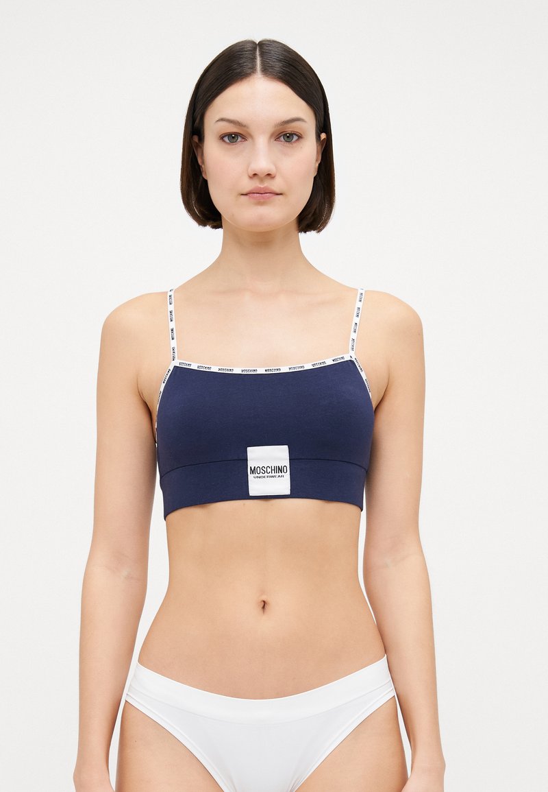 Bralette in cotone blu navy con spalline sottili e un'etichetta con logo bianco al centro. Texture liscia, design aderente e estetica minimalista.