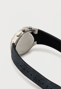 Ασήμι ρολόι χειρός με μαύρο υφασμάτινο λουράκι, που δείχνει το πίσω μέρος της θήκης και δύο πλαϊνά κουμπιά σε λευκό φόντο.